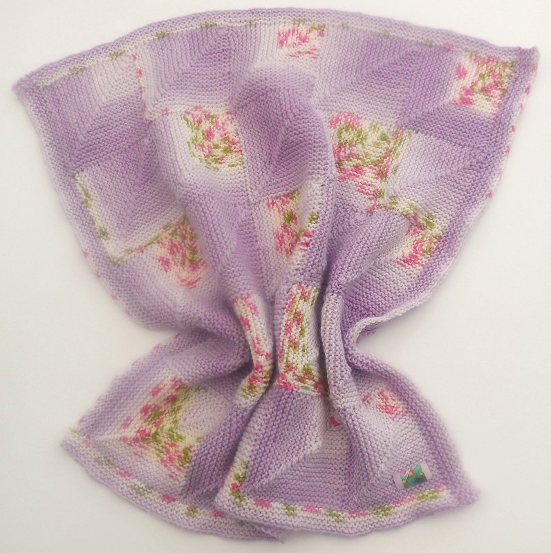 Gestrickte Babydecke in lila und weißen Quadraten, einige mit rosa und grünen Blumenmotiven.