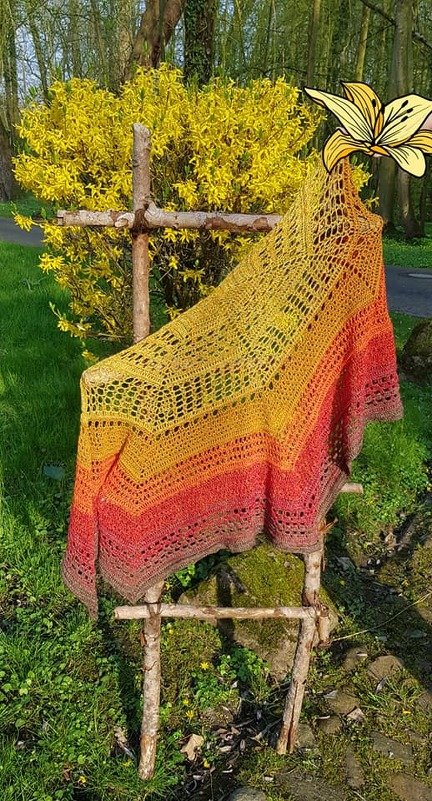 Gehäkeltes Dreieckstuch in Gelb, Orange und Rot auf rustikalem Holzständer vor gelben Blüten
