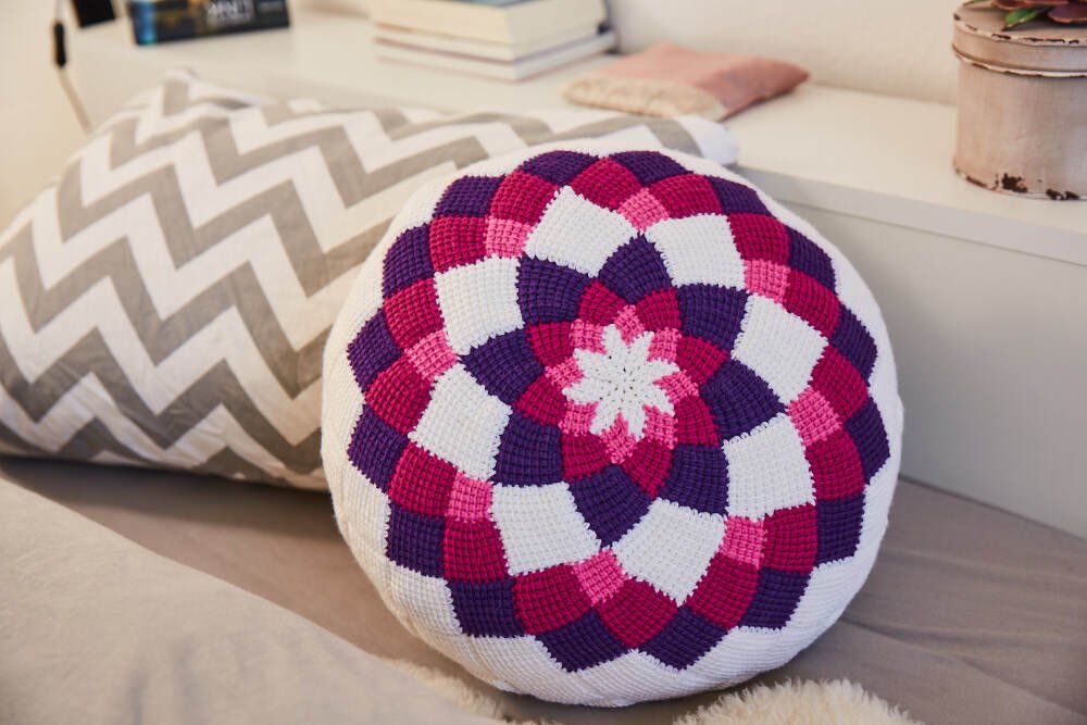 Rundes Mandala-Häkelkissen in Pink, Violett und Weiß neben einem Zickzack-Kissen