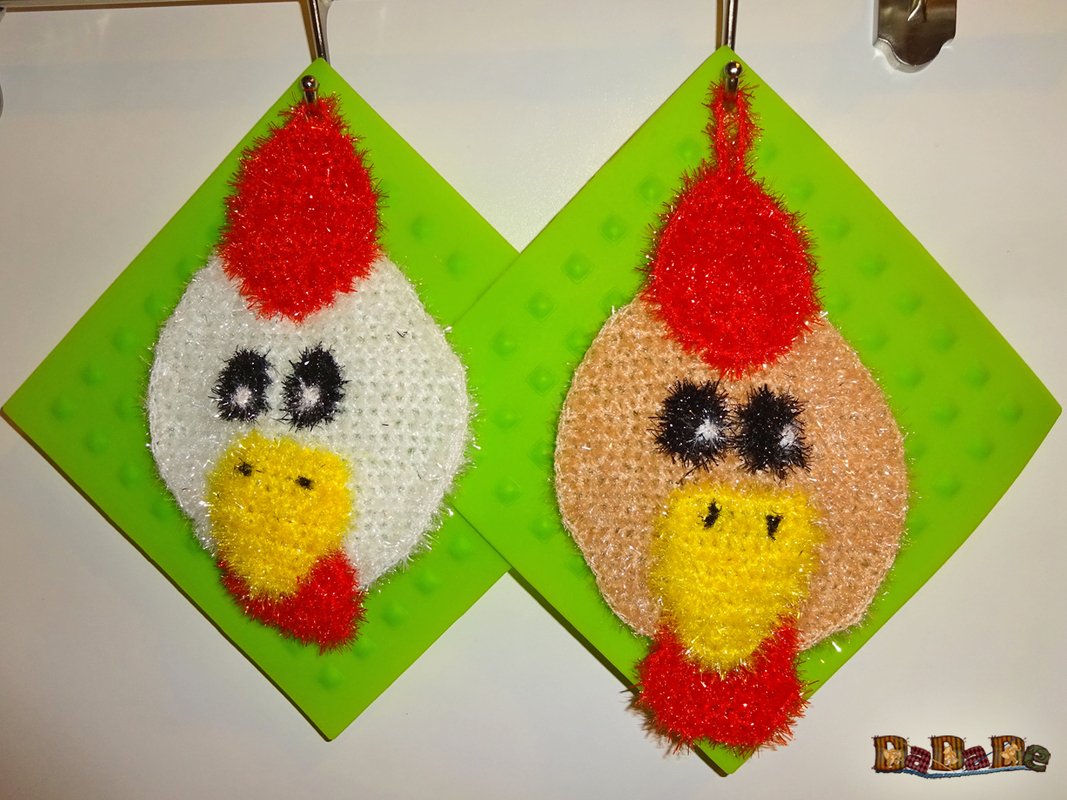 Zwei gehäkelte Hühnerkopf-Spüllappen, weiß und hellbraun, mit roten Kämmen und gelben Schnäbeln, an Haken aufgehängt