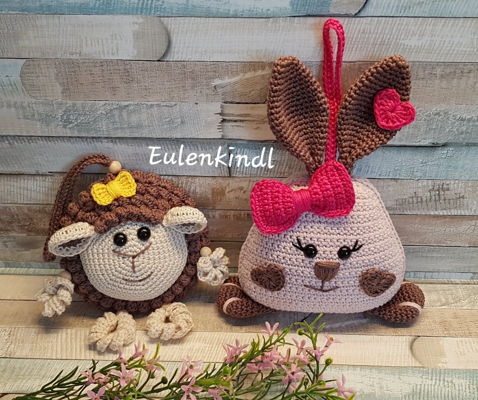 Zwei gehäkelte Amigurumi: ein hellrosa Osterhase mit pinker Schleife und Herz am Ohr neben einem braunen Schaf mit gelber Schleife vor Holzwand und rosa Blumen.
