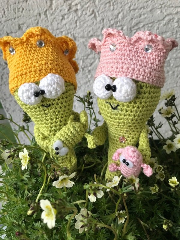 Zwei gehäkelte grüne Figuren mit gelber und rosa Krone und einer kleinen rosa Begleitfigur zwischen Pflanzen und weißen Blüten.