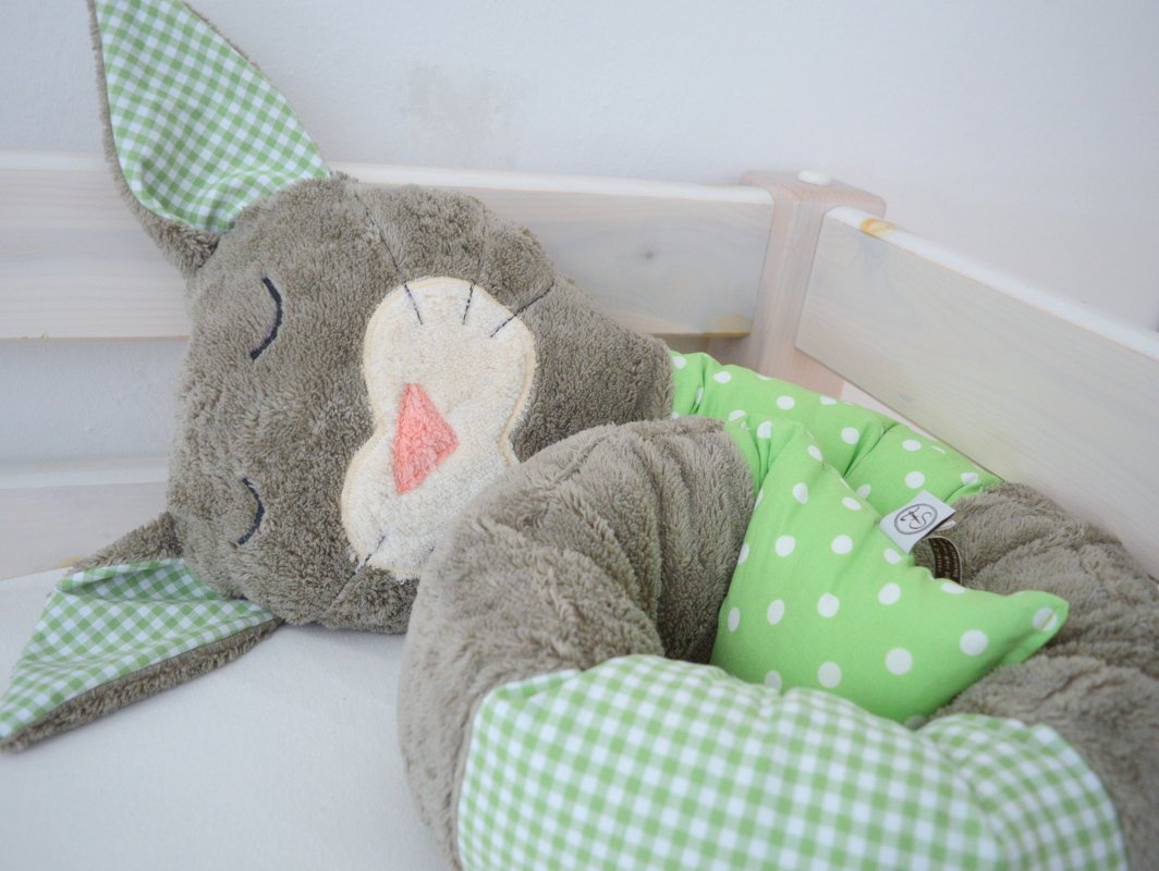 Graue Plüsch-Bettschlange in Hasenform mit grün-weiß karierten und gepunkteten Stoffeinsätzen in einem hellen Kinderbett.