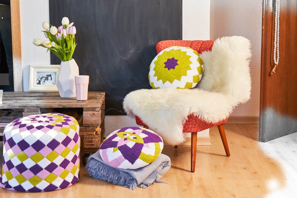 Wohnzimmerecke mit gehäkeltem Pouf, zwei runden Kissen, Sessel mit Schaffell und Palettentisch mit Blumen