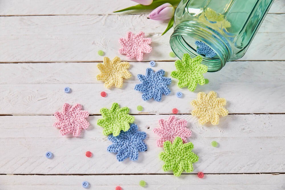 Mehrere gehäkelte kleine Blüten in Pastellfarben neben einem umgekippten Glas auf weißem Holzhintergrund, verstreute Knöpfe und eine Tulpe