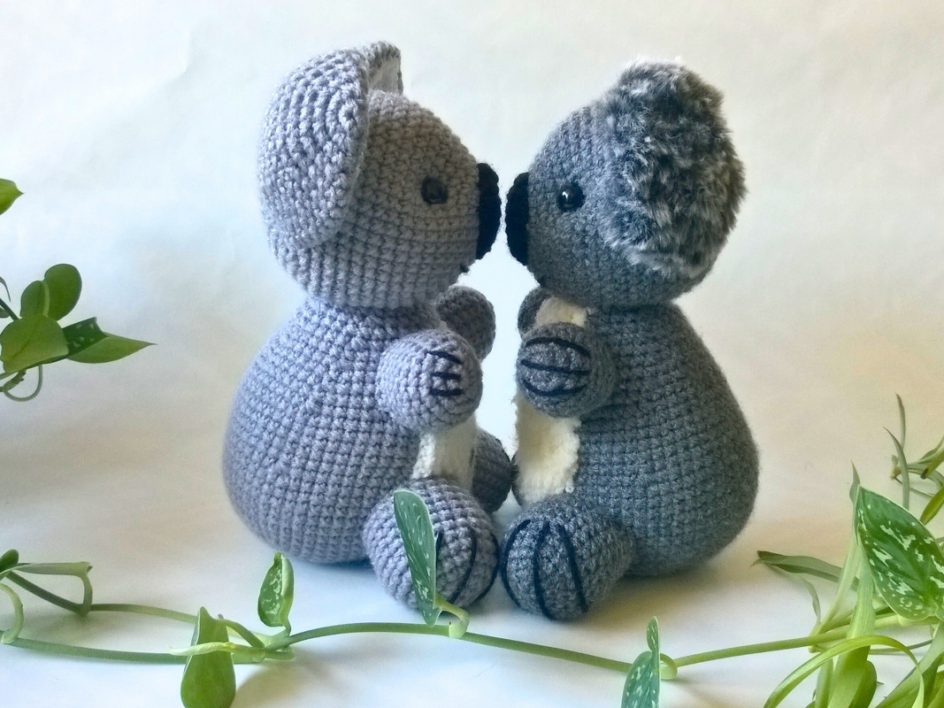 Zwei gehäkelte Amigurumi-Koalas in Hell- und Dunkelgrau, einer mit flauschigen Ohren, sitzen sich gegenüber, umgeben von grünen Pflanzen.