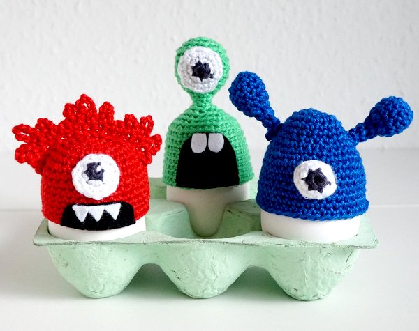 Drei gehäkelte Monster-Eierwärmer (rot, grün, blau) mit großen Augen und Filzdetails