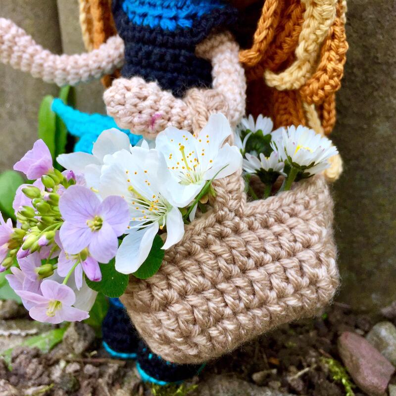 Nahaufnahme einer gehäkelten Figur mit kleinem beigen Korb voller weißer und violetter Frühlingsblumen