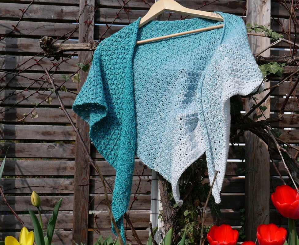 Gehäkeltes Dreieckstuch mit Farbverlauf von Türkis zu Weiß an einem Kleiderbügel vor einer Holzwand, rote und gelbe Tulpen im Vordergrund.
