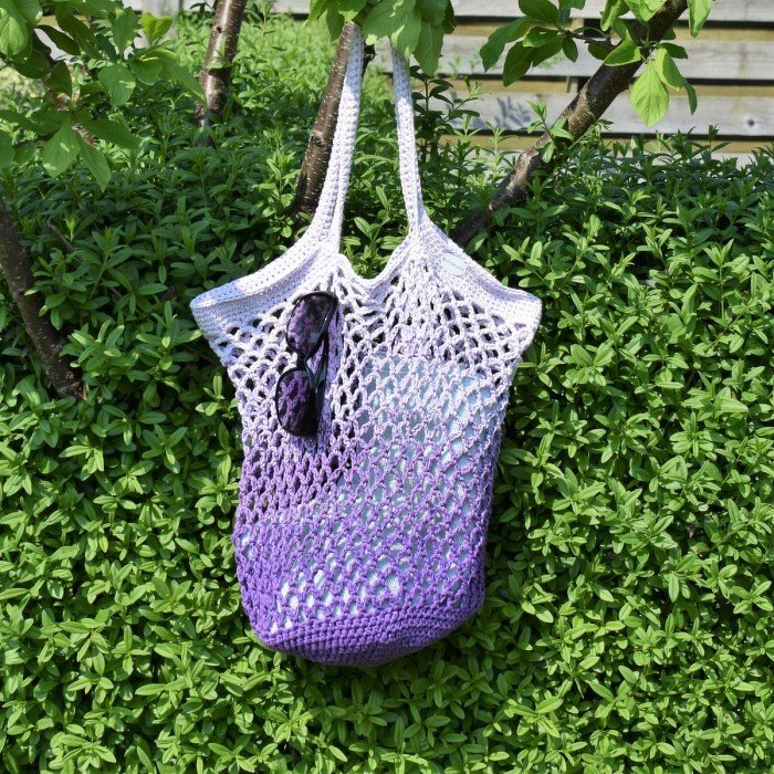 Gehäkelter Netzbeutel in Lila-Ombré hängt an Ast mit Sonnenbrille