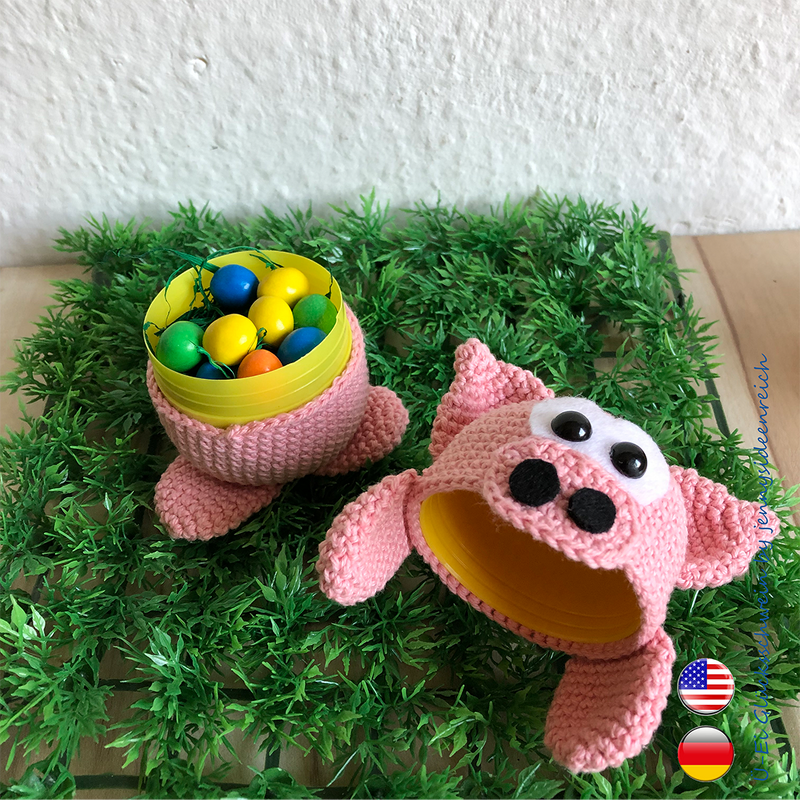 Two pink crocheted pig container covers on artificial grass, one holding colorful eggs.