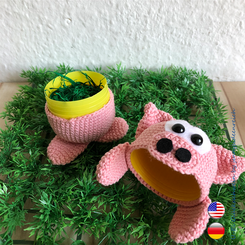 Nahansicht zweier rosa Häkel‑Schweinchen‑Hüllen mit gelben Innenkapseln auf grüner Deko