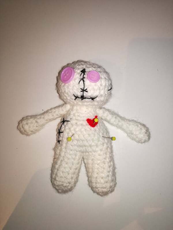 Kleine weiße Häkelpuppe mit rosa Knopfaugen, schwarzen Nähten, rotem Herz und Stecknadeln