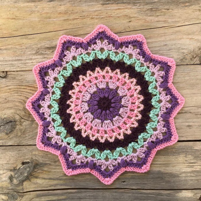 Round crocheted doily in pink, purple, mint green and dark brown with a star-shaped edge on a wooden surface.