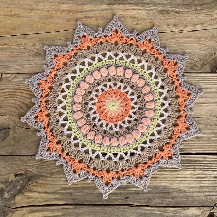 Round crocheted mandala doily with peach, beige, cream and pale green rings on a wooden plank background