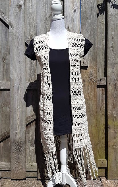 Long sleeveless beige crochet vest with openwork panels and long fringe on a mannequin in front of a wooden fence.