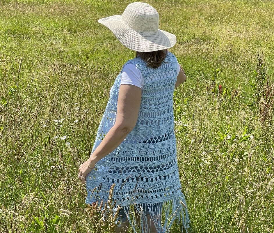 Rear view of a sleeveless long crochet vest with alternating openwork panels and fringe worn over a white t-shirt in a grassy field
