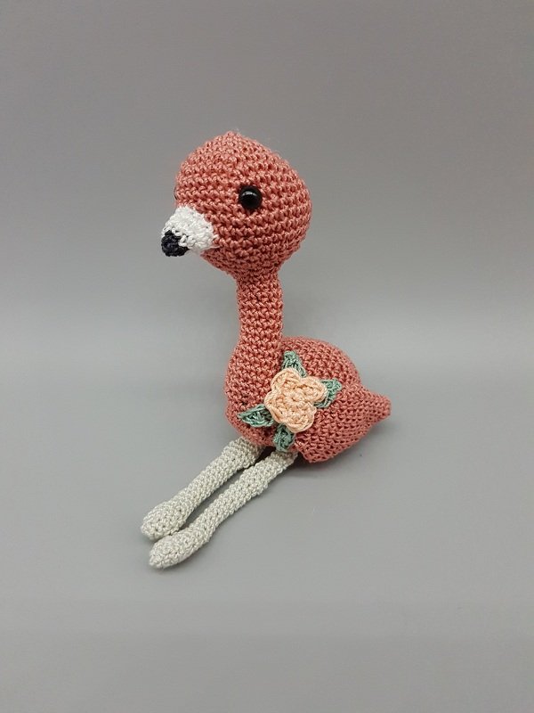 Gehäkeltes rosa Flamingo-Amigurumi mit langem Hals, hellen Beinen und Blütenapplikation am Körper