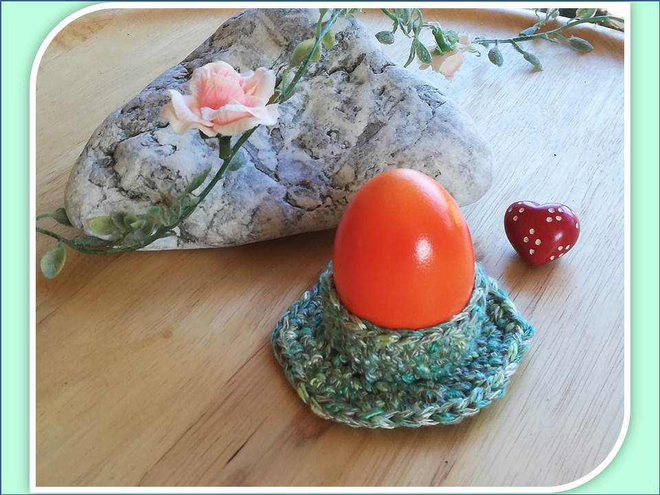 Gehäkelter türkisgrüner Eierbecher mit orangefarbenem Ei, Stein, rosa Blüte und rotem Herz auf Holz.
