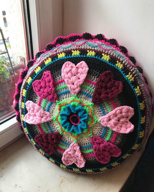 Rundes gehäkeltes Kissen mit zentralem Blumenmuster, applizierten Herzen in Pink- und Rosatönen, bunten Rändern und kleinen Zierperlen.