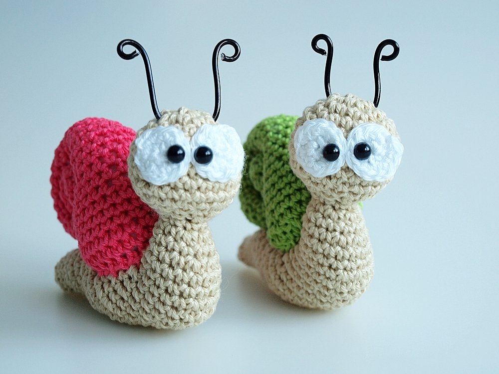Zwei gehäkelte Amigurumi-Schnecken mit rosa und grünem Gehäuse vor hellem Hintergrund