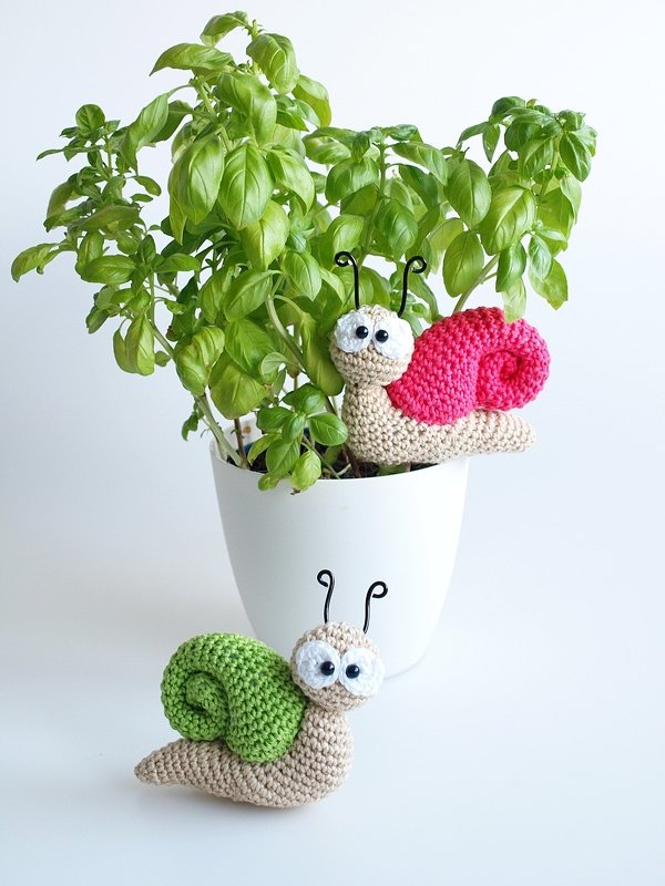 Zwei gehäkelte Amigurumi-Schnecken mit pinker und grüner Muschel neben einem Basilikumtopf