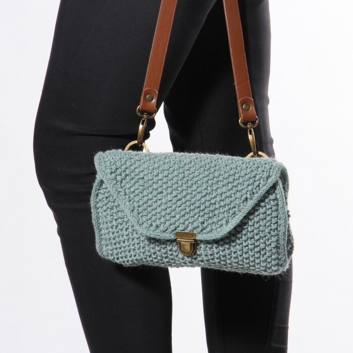 Light blue knitted shoulder bag with textured stitch, flap closure and brass turn lock, worn with black leggings.