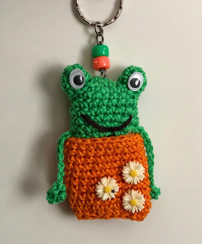 Gehäkelter Frosch-Schlüsselanhänger in Grün und Orange mit Wackelaugen, gesticktem Lächeln und drei kleinen Blüten auf dem Bauch
