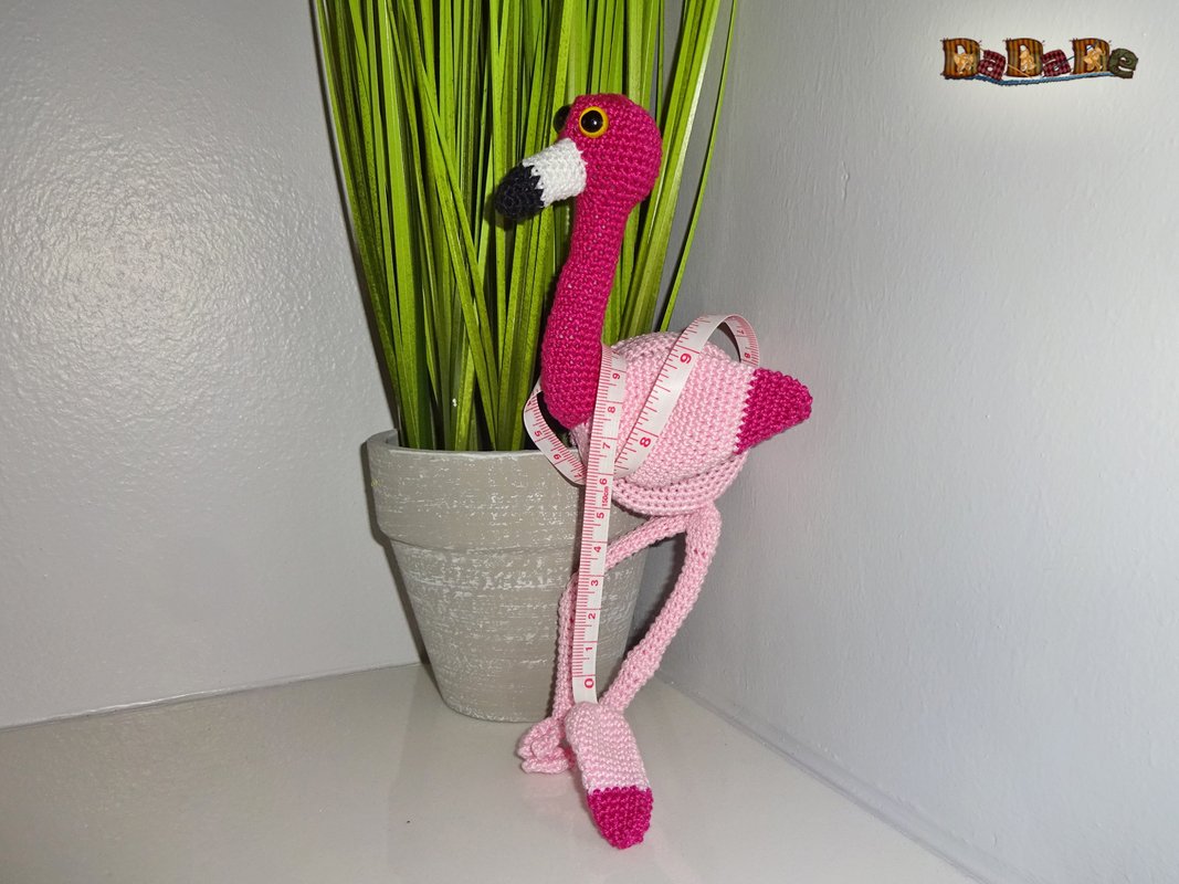 Gehäkelter pinker Flamingo mit locker um den Körper geschlungenem Maßband neben einer Topfpflanze auf heller Ablage.
