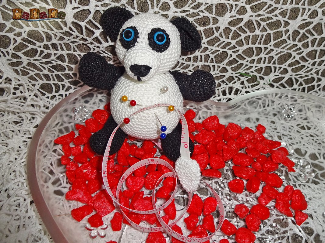 Gehäkeltes Panda-Nadelkissen mit blauen Augen, bunten Stecknadeln und Maßband in einer herzförmigen Schale auf roten Dekosteinen.
