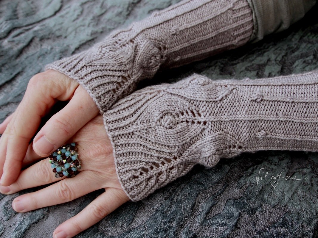Pair of light brown knitted fingerless gloves with cable and lace stitch details worn on hands, resting on a textured fabric surface.