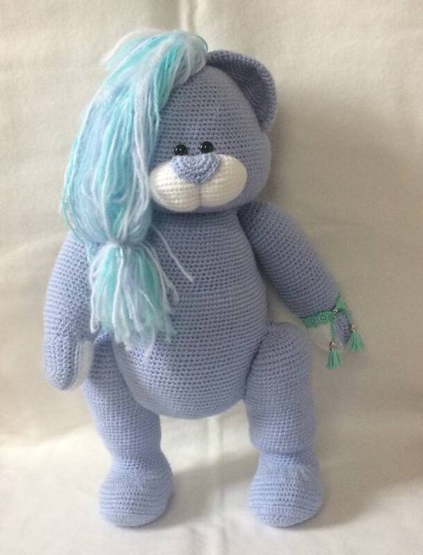 Gehäkelter fliederfarbener Amigurumi-Teddy mit türkis-weißem Garnzopf, weißer Schnauze und grünem Armband