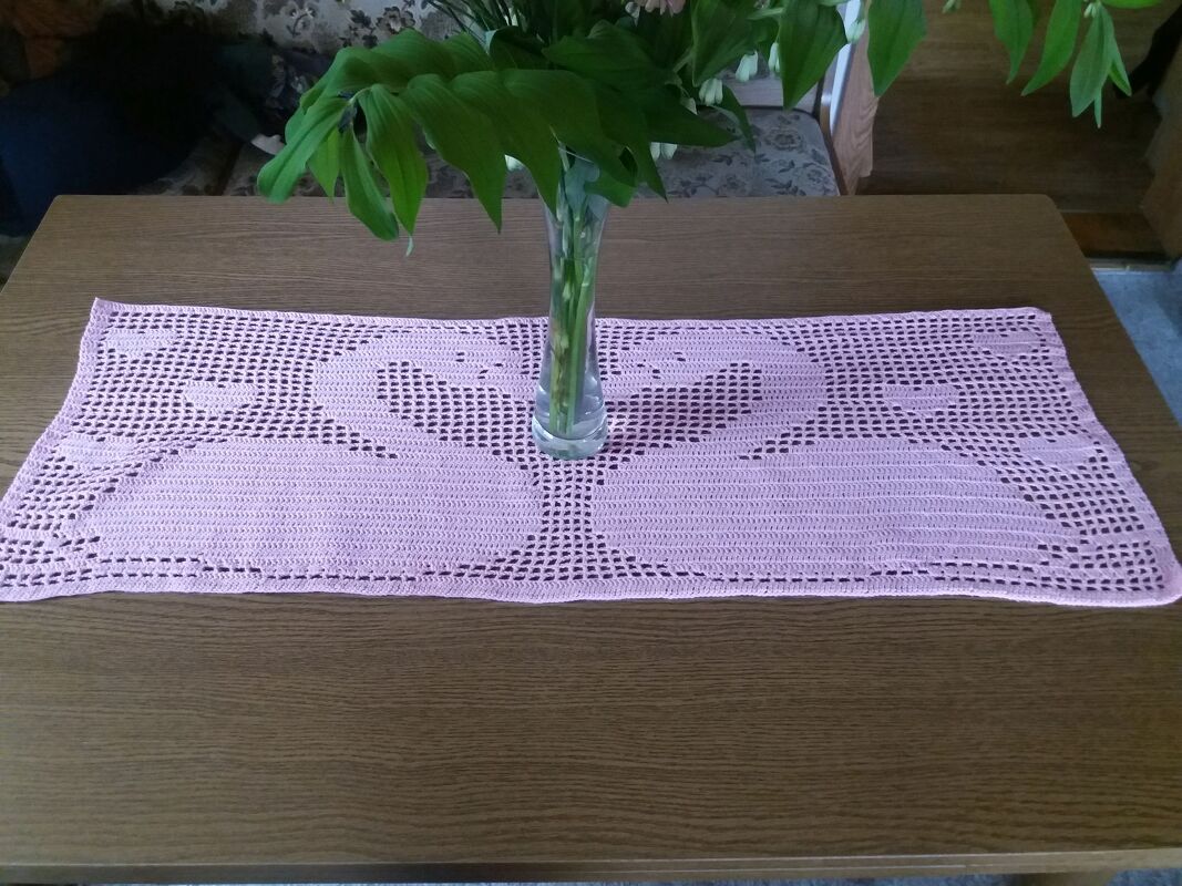 Zartrosa gehäkelter Tischläufer mit zwei Schwänen auf einem Holztisch