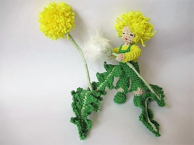 Gehäkeltes Amigurumi einer Löwenzahn‑Fee mit gelbem Haar und grüner Blätterkleidung neben gelber Blüte und weißer Pusteblume auf hellem Hintergrund
