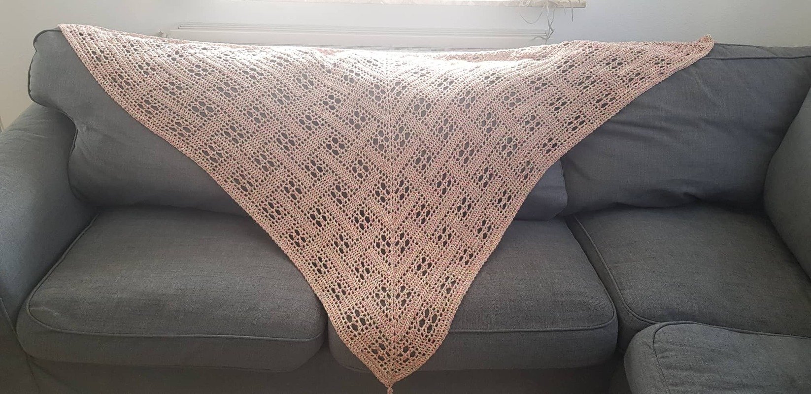 Pastellrosa gehäkeltes Dreieckstuch mit Spitzen- und Lochmuster auf grauem Sofa