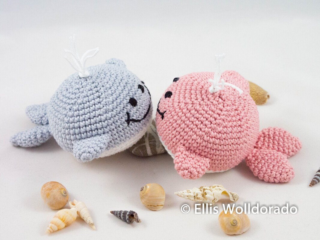 Zwei gehäkelte Amigurumi-Wale in Pastellblau und Rosa, liegend neben Muscheln
