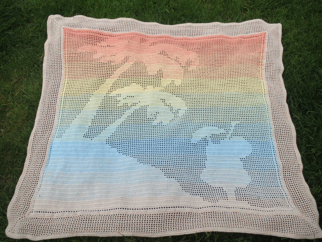 Häkeldecke im Filethäkelmuster mit Pferdemotiven und Rosa-Gelb-Blau-Farbverlauf auf grünem Rasen