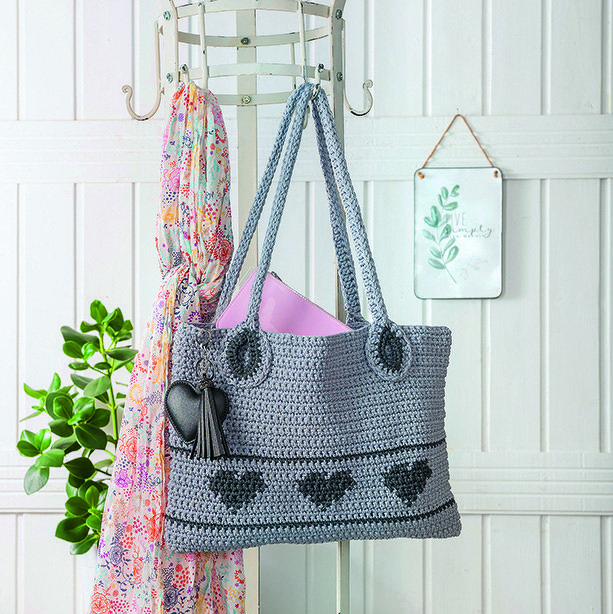 Gehäkelte graue Henkeltasche mit dunkelgrauen Herzen und herzförmigem Anhänger, an Garderobenhaken hängend