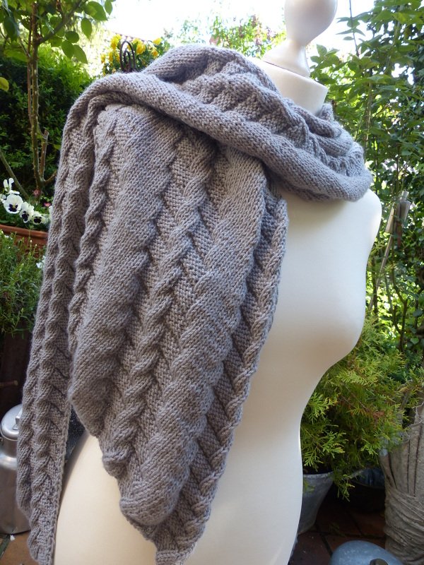 Gestrickter grauer Schal mit Zopfmuster auf einer Schaufensterpuppe, im Hintergrund Pflanzen