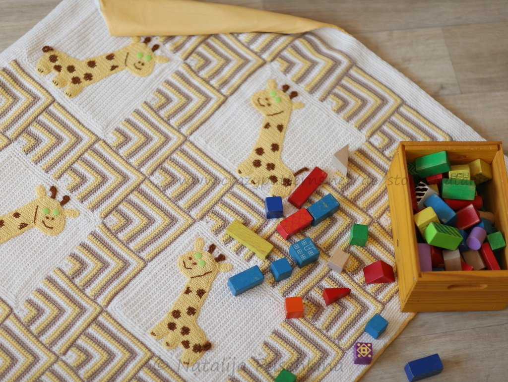 Handgehäkelte Babydecke in Weiß, Gelb und Braun mit Giraffen-Applikationen und geometrischem Muster, umgeben von Holzbausteinen.