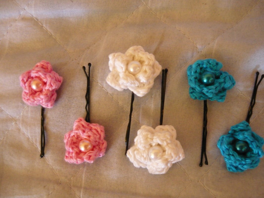 Sechs gehäkelte Blumen-Haarnadeln: jeweils zwei in Rosa, Weiß und Türkis, mit Perlen in der Mitte auf gestepptem Stoff.