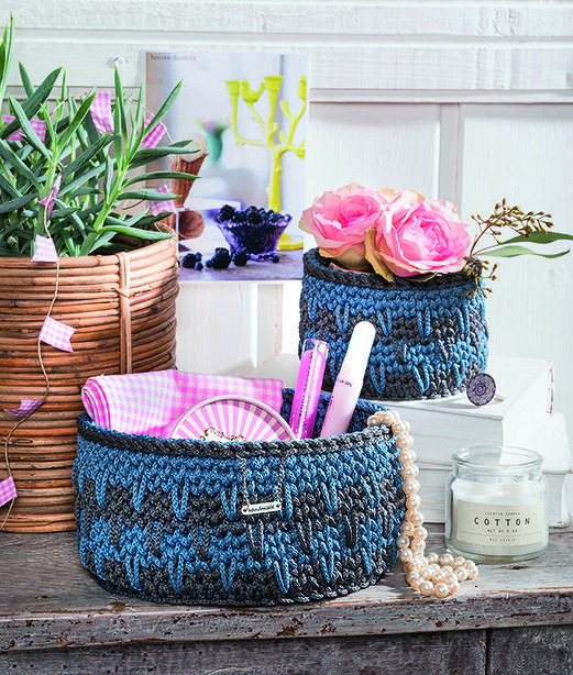 Zwei gehäkelte Körbe in Blau und Grau auf einer Holzoberfläche, einer gefüllt mit Kosmetikartikeln, der andere mit Rosen; daneben Topfpflanze und Kerze.
