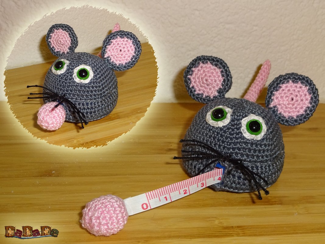Gehäkelte dunkelgraue Maus mit rosa Ohren und Nase; aus ihrem Maul wird ein weißes, ausziehbares Rollmaßband mit rosa Häkelkugel gezogen.