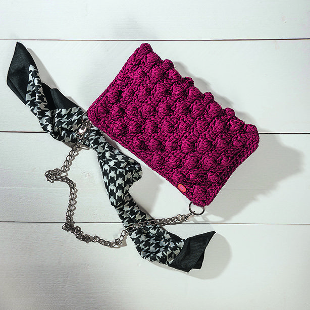 Bordeauxrote gehäkelte Clutch mit Noppenmuster, silberner Gliederkette und schwarz-weißem Tuch auf hellem Holzhintergrund.