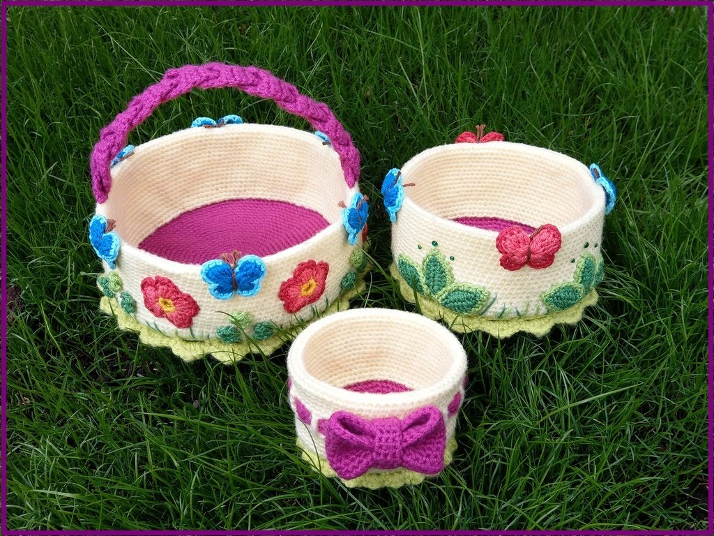 Drei gehäkelte Körbe in Creme und Pink mit blauen Schmetterlingen und bunten Blumen auf Gras