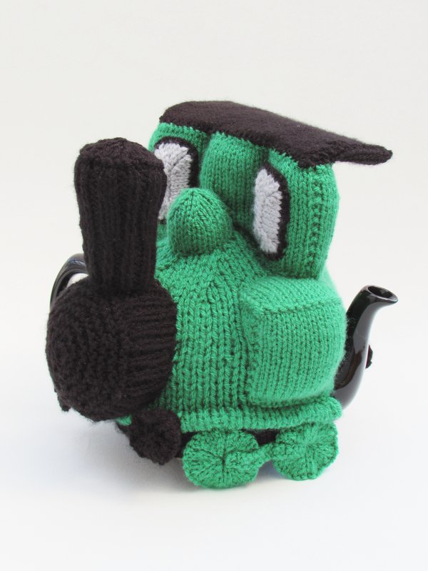Green knitted train teapot cozy with black smokestack covering a teapot, showing spout and handle