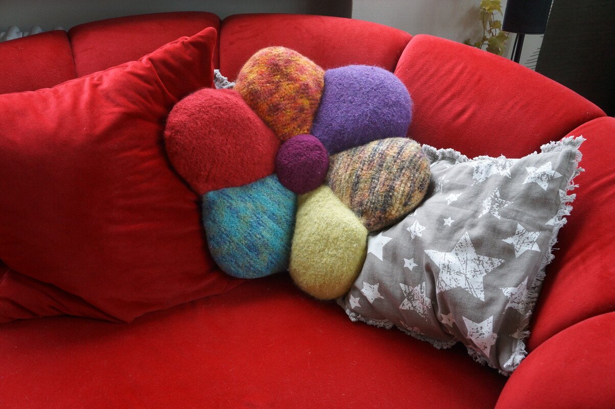 Multicolored flower-shaped knitted and felted cushion with seven petals on a red sofa beside a grey star-print pillow