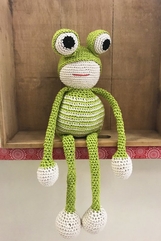 Gehäkelter Frosch Amigurumi in Grün-Weiß mit großen Augen und langen Gliedmaßen, sitzend auf einem Holzregal.