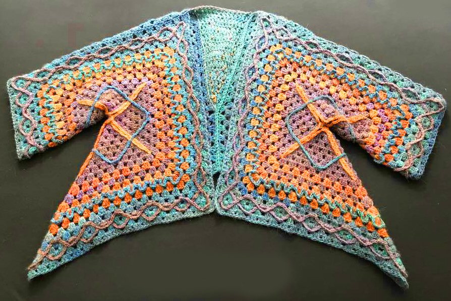 Bunte gehäkelte Damenjacke im Granny-Square-Muster mit wellenförmiger Saumkante, offen auf dunklem Untergrund liegend.