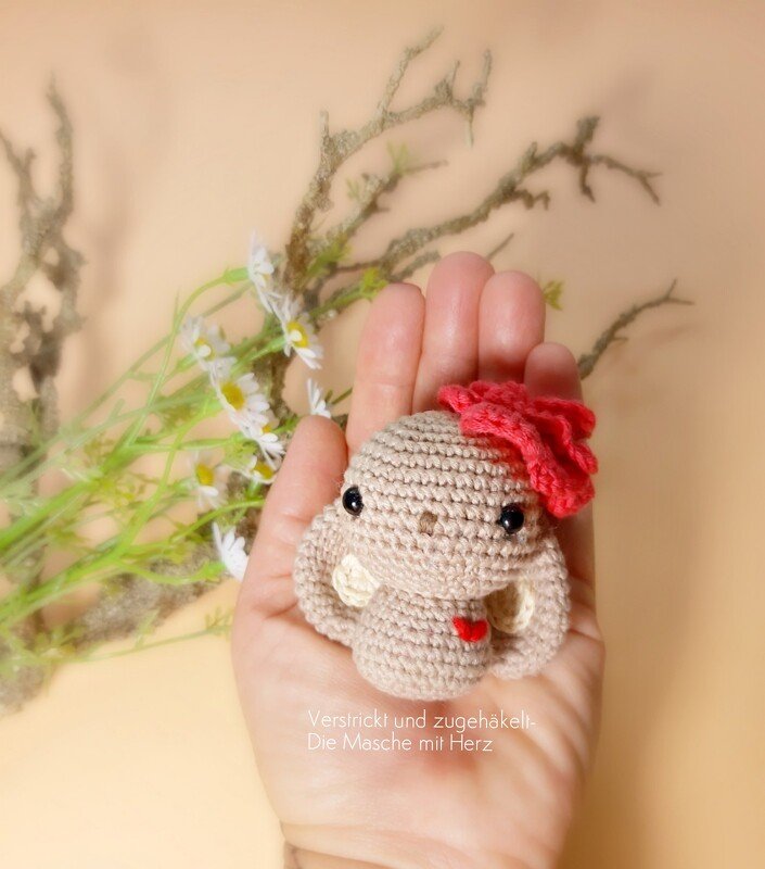 Kleiner beiger Amigurumi-Hase mit roter Blume am Kopf und kleinem roten Herz, in einer Hand gehalten